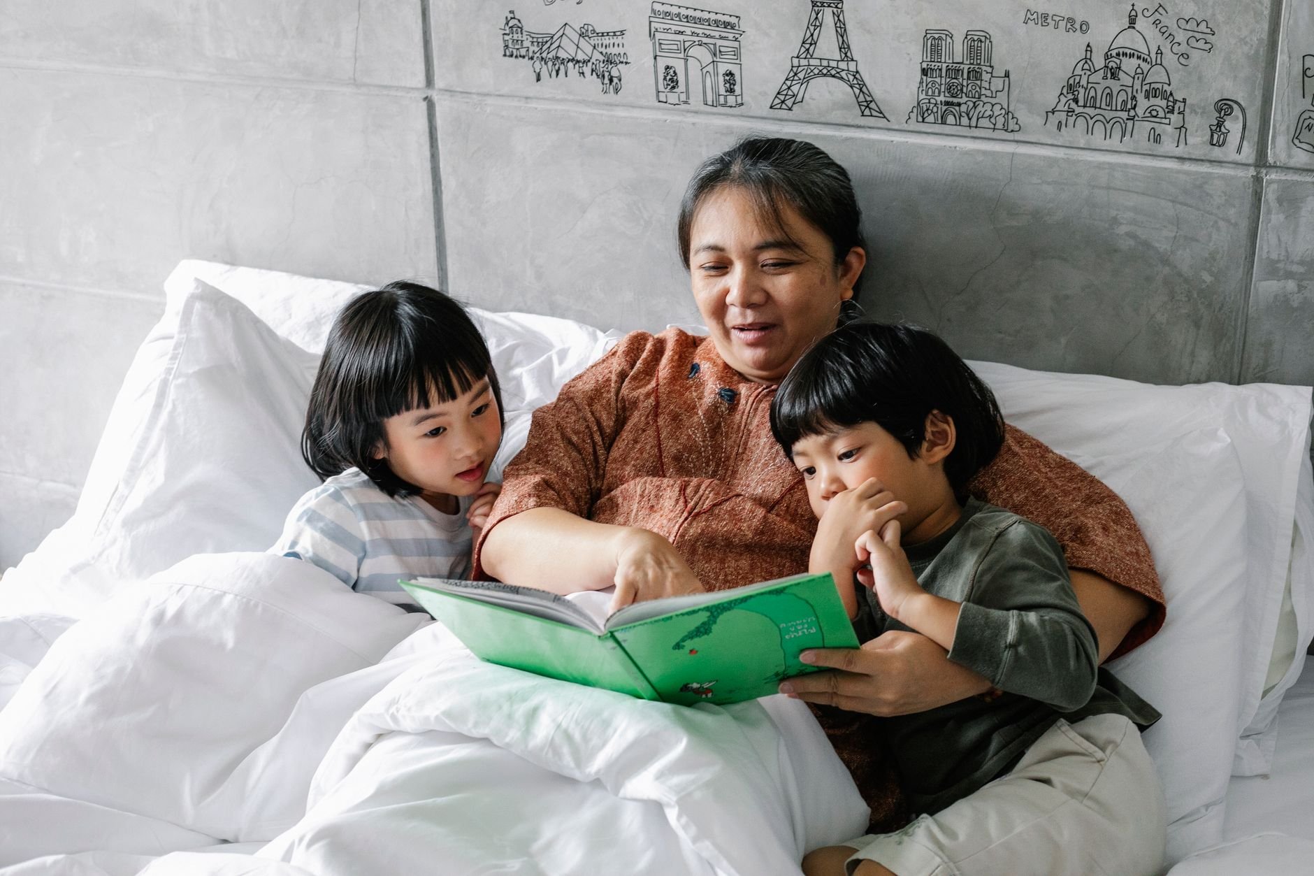 From above, a grandmother reads a fairytale to young children lying on a soft bed — a warm, multigenerational story moment.